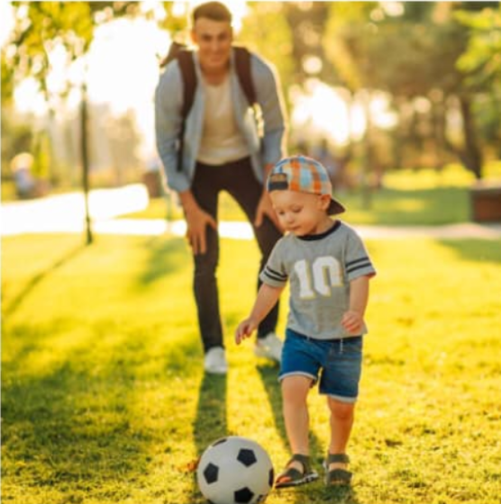 Un padre sonríe mientras observa a su hijo pequeño jugar con una pelota de fútbol en un parque, con la luz dorada del atardecer detrás de ellos.
